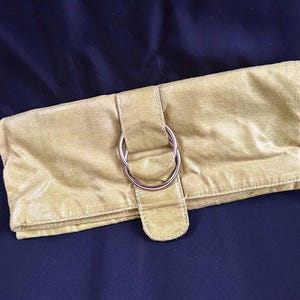 May include: A light olive green clutch with a silver-colored metal ring closure. The clutch is made of a soft, suede-like material and has a rectangular shape. The background is a dark blue fabric.