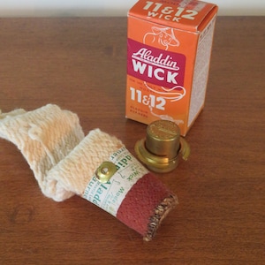 May include: A red and white box with the text "Aladdin Wick 11 & 12" on it. The box is next to a white and brown wick with a gold metal cap. The wick is labeled "Aladdin Wick".