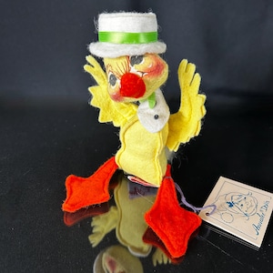 May include: A vintage Analee Dolls duck figurine. The duck is yellow with orange feet and a red beak. It wears a white top hat with a green band and a white scarf. The doll has black eyes and a tag attached with a purple string.