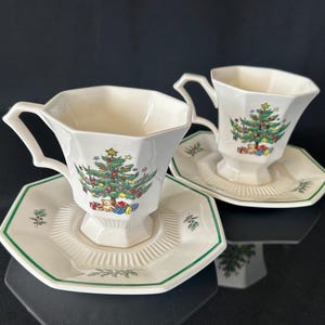 May include: Two white teacups and saucers with a Christmas tree design. The cups have a unique angular shape and a handle. The saucers are octagonal with a green border. The Christmas tree is decorated with ornaments and presents.
