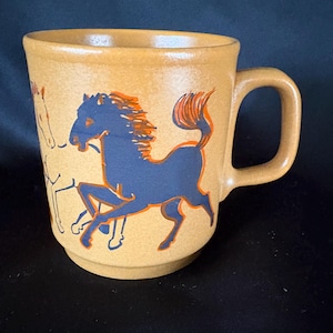 May include: A tan ceramic mug featuring a dark blue graphic of a galloping horse with orange accents. The mug has a cylindrical body with a stepped base and a sturdy side handle. The design shows a horse in motion with a flowing mane and tail.