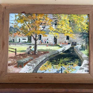 May include: A framed painting of a waterside scene with a stone building, a tree with yellow leaves, and a canal. The artwork features figures, a wooden fence, and a stone bench. The frame is made of wood.