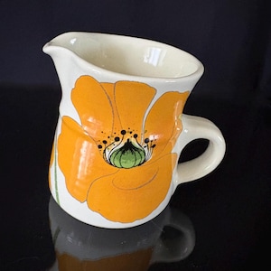May include: Cream-colored ceramic pitcher with a large orange poppy flower design. The flower has black and green details. The pitcher has a handle and a spout for pouring. A decorative floral accent for the kitchen.