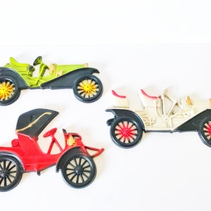 Puede incluir: Tres coches de juguete vintage en diferentes colores. Un coche verde, un coche blanco y un coche rojo. Cada coche tiene ruedas negras y detalles dorados.