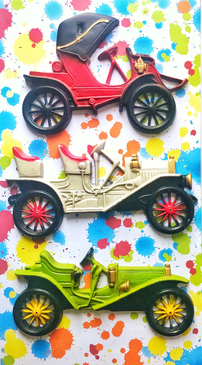 Puede incluir: Tres coches de juguete vintage en rojo, blanco y verde. Los coches est&aacute;n hechos de metal y tienen un dise&ntilde;o detallado. Est&aacute;n colocados sobre un fondo blanco con salpicaduras de pintura de colores.