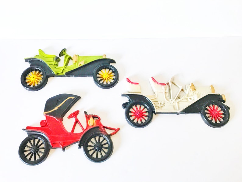 Puede incluir: Tres coches de juguete vintage en diferentes colores. Un coche verde, un coche blanco y un coche rojo. Cada coche tiene ruedas negras y detalles dorados.