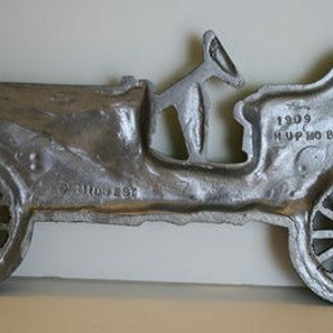 Puede incluir: Una escultura de pared de metal plateado de un autom&oacute;vil Hupmobile de 1909. La escultura est&aacute; detallada con las ruedas, la carrocer&iacute;a y un asiento de conductor. El texto "1909 HUPMOBILE" es visible en la parte posterior de la escultura.