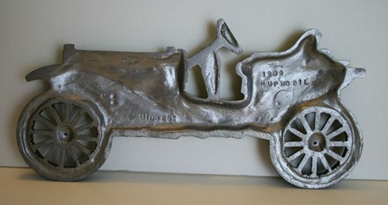 Puede incluir: Una escultura de pared de metal plateado de un autom&oacute;vil Hupmobile de 1909. La escultura est&aacute; detallada con las ruedas, la carrocer&iacute;a y un asiento de conductor. El texto "1909 HUPMOBILE" es visible en la parte posterior de la escultura.