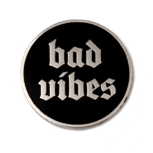 May include: A black and silver circular pin with the text "bad vibes" in a gothic font.