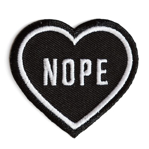 May include: Black heart-shaped patch with white stitching around the edges. The word "NOPE" is embroidered in white on the patch.