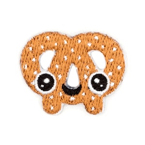 May include: A brown embroidered patch shaped like a pretzel with a smiling face. The pretzel has white dots on it.