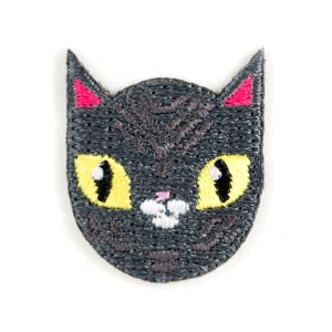 May include: Embroidered patch featuring a stylized cat face. The cat has dark gray fur, yellow eyes with black pupils, pink inner ears, and a small pink nose. The patch is designed with a textured, stitched appearance, suitable for application on fabric items.