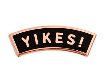 May include: A rose gold-colored metal pin with a black enamel background. The pin is shaped like an arch and says "YIKES!" in white letters.