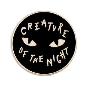 May include: Black enamel pin with a white outline of the text "Creature of the Night" and two white eyes.