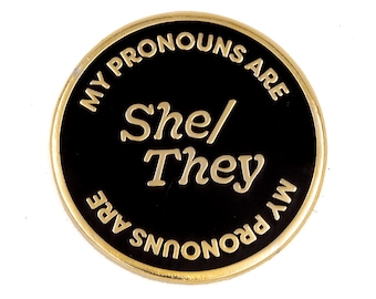 May include: Gold-colored pin with a black background. The text "My pronouns are She/They My pronouns are" is written in a circle around the edge of the pin. The words "She/They" are written in larger font in the center of the pin.