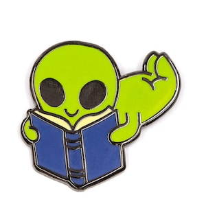 May include: A green alien with a big smile is reading a blue book. The alien has one arm raised in the air, with a closed fist.