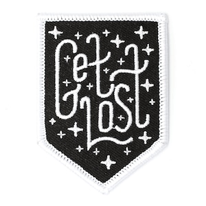 May include: Black and white embroidered patch with a white outline. The patch is shaped like a shield and features the text "Get Lost" in a stylized font. The background is black with white stars.