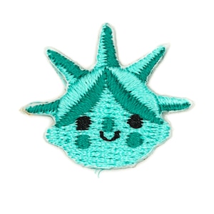 May include: Embroidered patch featuring a cartoon version of the Statue of Liberty. The patch is teal with dark green accents and black eyes and a smile. The patch has a white border and is likely for clothing or accessories.