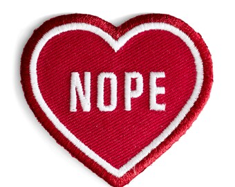 May include: A heart-shaped patch with a red background and white embroidered text that reads "NOPE." The patch has a white border with a red outline. The patch is likely for clothing or accessories.