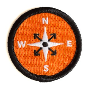 May include: Orange fabric compass patch with a white compass rose and black lettering. The compass points are labeled N, S, E, and W.