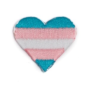 May include: A heart-shaped patch with the transgender pride flag colors: light blue, pink, and white stripes.