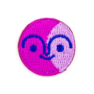 May include: A pink and purple embroidered patch with a smiling face. The face is split in half, with one side pink and the other purple. The eyes are blue.