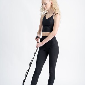May include: A person wearing black leggings and a black sports bra uses a blue and black ankle strap to stretch their foot.