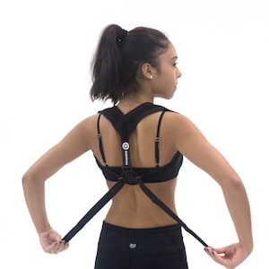 May include: A person wearing a black posture corrector with adjustable straps. The corrector has the text "Kinetic" printed on it.