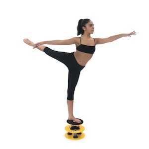 May include: A person in black leggings and a black sports bra stands on a yellow and black balance disc. The disc has a spring mechanism and is stacked on top of another similar disc.