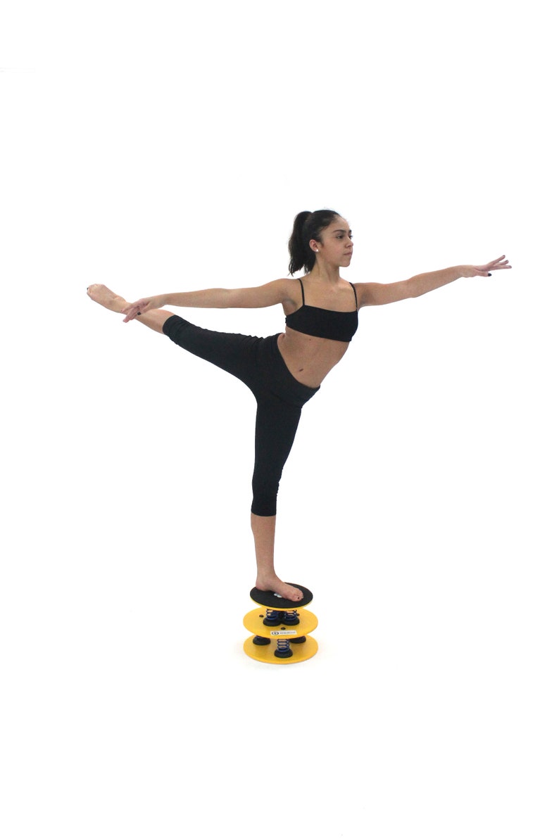 May include: A person in black leggings and a black sports bra stands on a yellow and black balance disc. The disc has a spring mechanism and is stacked on top of another similar disc.