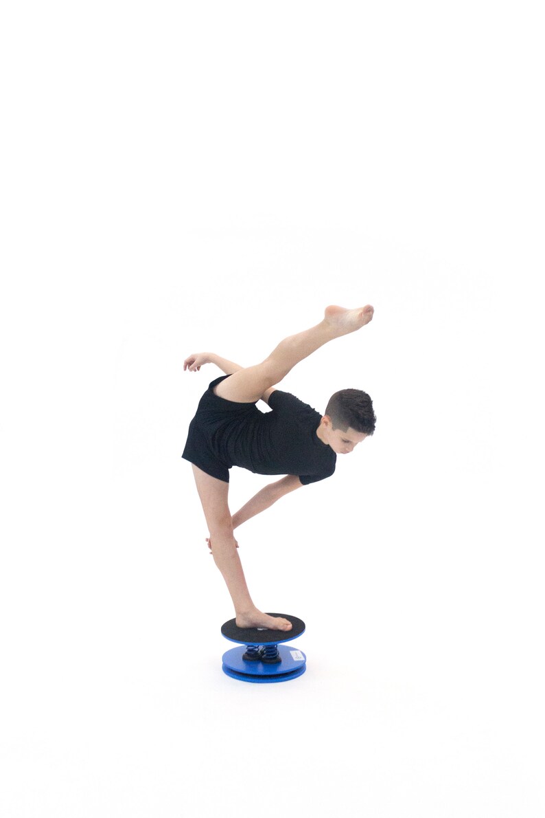 Puede incluir: Una persona con ropa deportiva negra se equilibra sobre una tabla de equilibrio azul y negra. La tabla tiene una parte superior e inferior circulares conectadas por un resorte. La persona est&aacute; realizando un ejercicio de equilibrio.