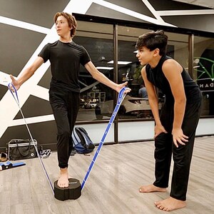 May include: Two people are stretching with a blue resistance band. The person on the left is standing on a black cylinder and holding the band with both hands. The person on the right is standing with their feet apart and holding the band with one hand.