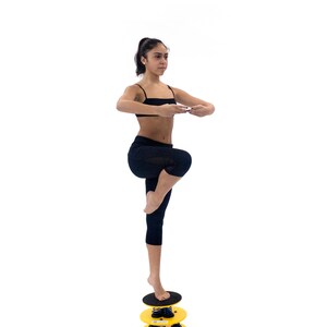 May include: A person standing on a yellow and black spring-loaded balance disc. The disc has three tiers and is designed for balance and core strengthening exercises.