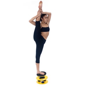 May include: A woman in black leggings and a black sports bra is balancing on a yellow and black balance disc. She is holding her leg up in the air, demonstrating a yoga pose.
