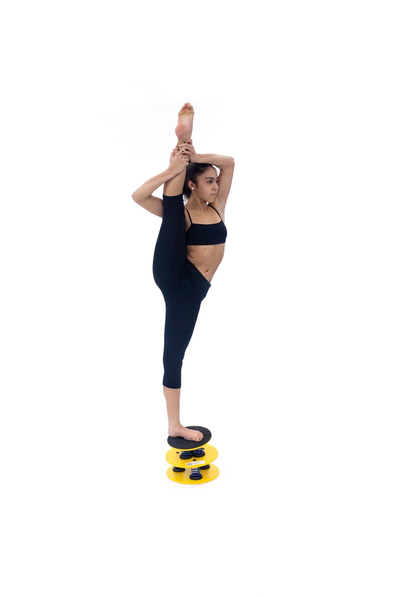 May include: A woman in black leggings and a black sports bra is balancing on a yellow and black balance disc. She is holding her leg up in the air, demonstrating a yoga pose.