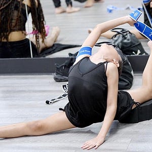 May include: A young person wearing a black tank top and blue resistance bands stretches on a black foam block. The person is doing a split with one leg extended forward and the other leg bent behind them.
