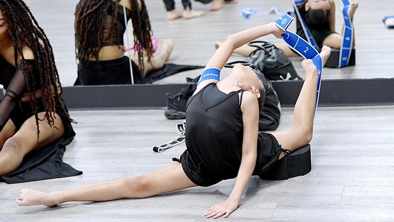 May include: A young person wearing a black tank top and blue resistance bands stretches on a black foam block. The person is doing a split with one leg extended forward and the other leg bent behind them.