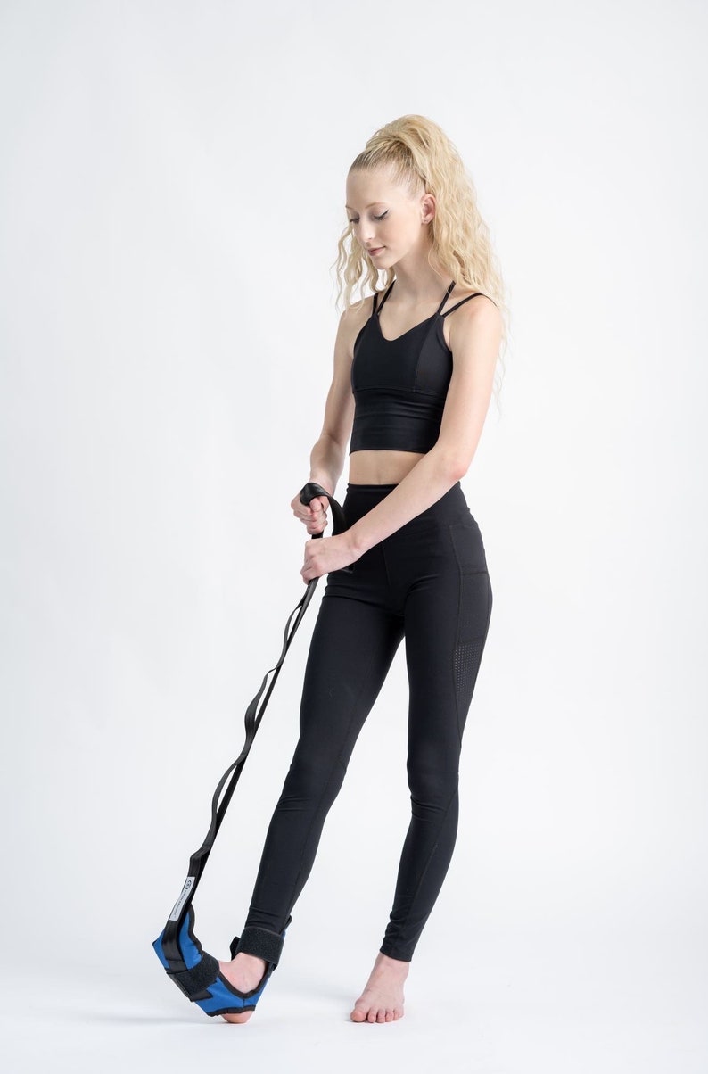 May include: A person wearing black leggings and a black sports bra uses a blue and black ankle strap to stretch their foot.
