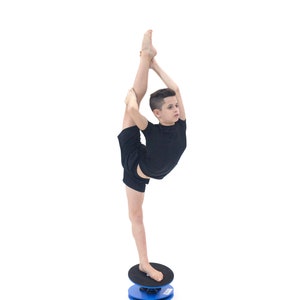 Puede incluir: Una persona con ropa deportiva negra se equilibra sobre una tabla de equilibrio azul y negra, realizando una apertura vertical. La tabla es redonda, con la parte superior negra y la base azul. El fondo es blanco.