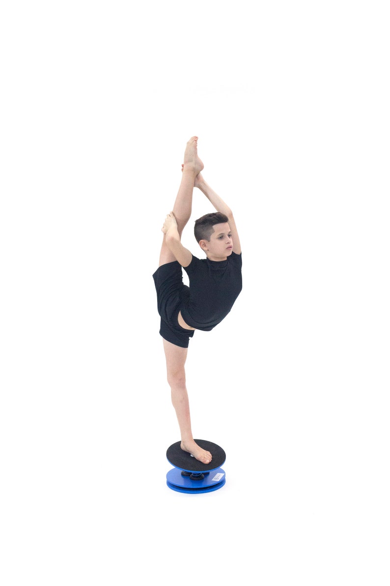 Puede incluir: Una persona con ropa deportiva negra se equilibra sobre una tabla de equilibrio azul y negra, realizando una apertura vertical. La tabla es redonda, con la parte superior negra y la base azul. El fondo es blanco.