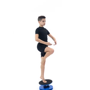 Puede incluir: Una persona con un atuendo negro se equilibra sobre una tabla de equilibrio azul y negra. La tabla de equilibrio tiene una base redonda azul y una parte superior negra texturizada. La persona est&aacute; descalza y de pie sobre una pierna, demostrando equilibrio.