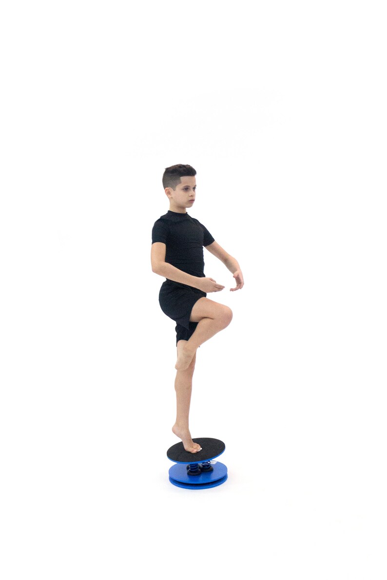 Puede incluir: Una persona con un atuendo negro se equilibra sobre una tabla de equilibrio azul y negra. La tabla de equilibrio tiene una base redonda azul y una parte superior negra texturizada. La persona est&aacute; descalza y de pie sobre una pierna, demostrando equilibrio.