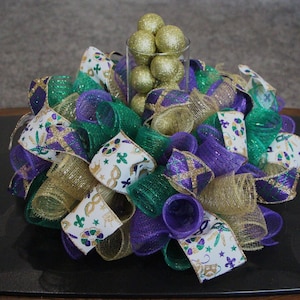May include: A Mardi Gras themed centerpiece with gold, purple, and green mesh ribbon, gold glitter balls, and a clear glass vase.
