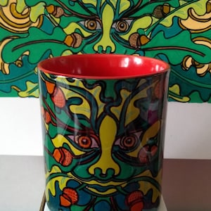 May include: A red and green ceramic mug with a green man face design. The face is surrounded by green leaves and acorns. The mug is on a white surface.