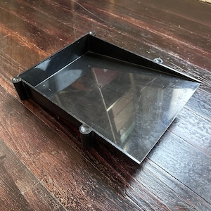 May include: A black plastic tray with a sloped front edge, resting on a dark wooden surface. The tray has a rectangular shape with rounded corners and is designed for collecting or sorting items. The tray is empty.