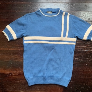 May include: A light blue knit short-sleeve shirt with cream-colored stripes. The shirt has a crew neck and ribbed cuffs and hem. The stripes are on the sleeves and across the chest. The shirt is made of a soft, knitted material.