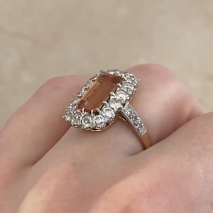 May include: A gold ring featuring a large, rectangular, peach-colored gemstone surrounded by a halo of smaller, clear gemstones. The ring's band is also adorned with small, clear gemstones.