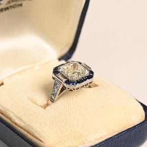 Anillo con diamante talla Asscher de 3 quilates, certificado por el GIA. Engaste biselado. Detalle de zafiro talla francesa tipo halo. Elaborado a mano en platino.