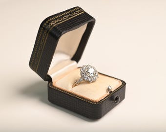 May include: A diamond engagement ring in an open black leatherette box. The ring has a central diamond surrounded by smaller stones, set on a gold band. The box is trimmed with gold and has a light beige interior. The box is open.