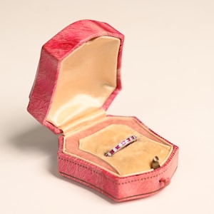 May include: An open, pink, hexagonal jewelry box with a ring inside. The ring is silver-toned with a row of small, pink gemstones. The box has a light tan interior and a small clasp.